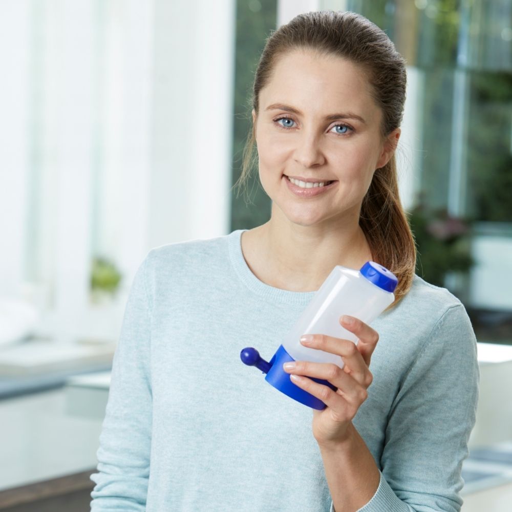 Une femme tient une douche nasale avec un couvercle et une base bleus. Elle sourit à la caméra.