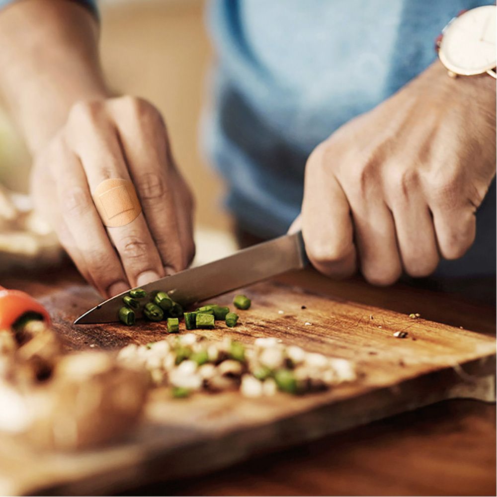 Personne coupant des légumes avec un couteau sur une planche à découper. Un pansement sur le doigt.