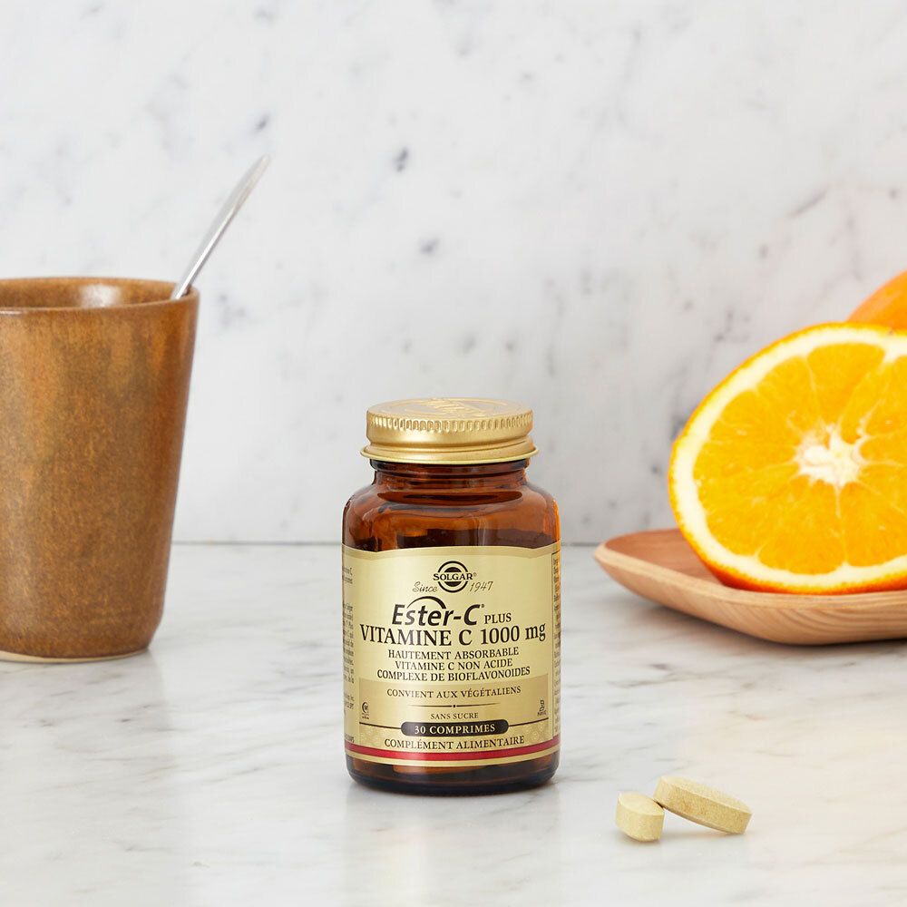 Flacon avec comprimés, tasse et fruits. Nom du produit: Ester-C Plus 1000 mg. Sur une table.