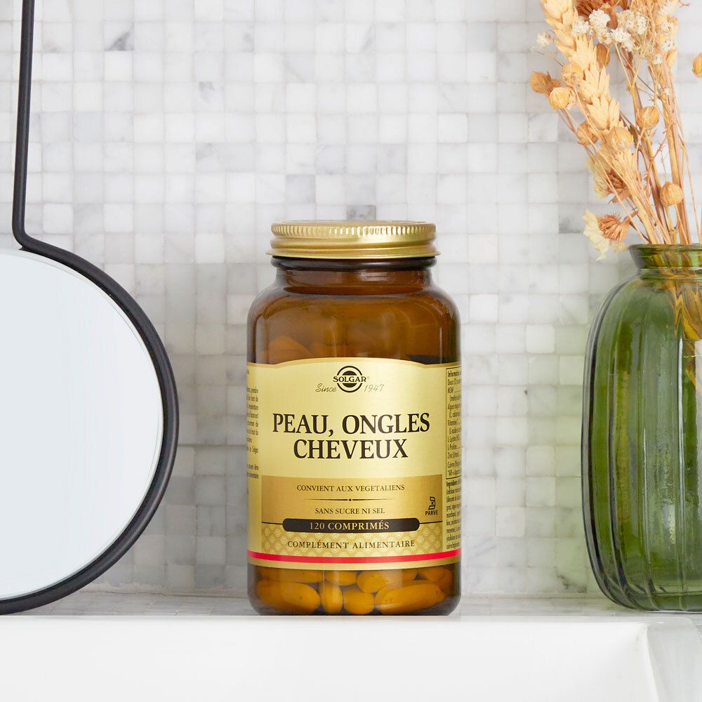 Flacon en verre avec bouchon doré. Inscription « Peau, Ongles, Cheveux ». Posé sur une surface blanche.