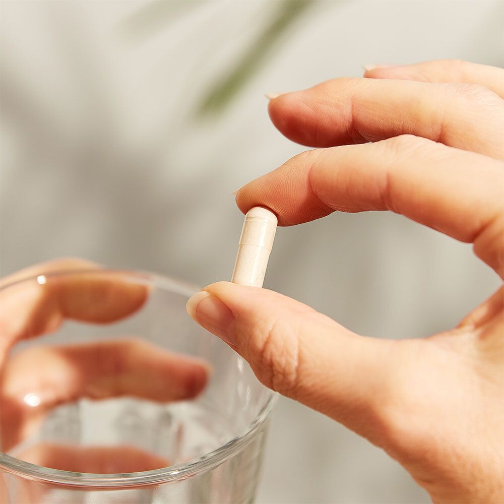 Main tenant une gélule au-dessus d'un verre d'eau. Peau claire, arrière-plan flou.