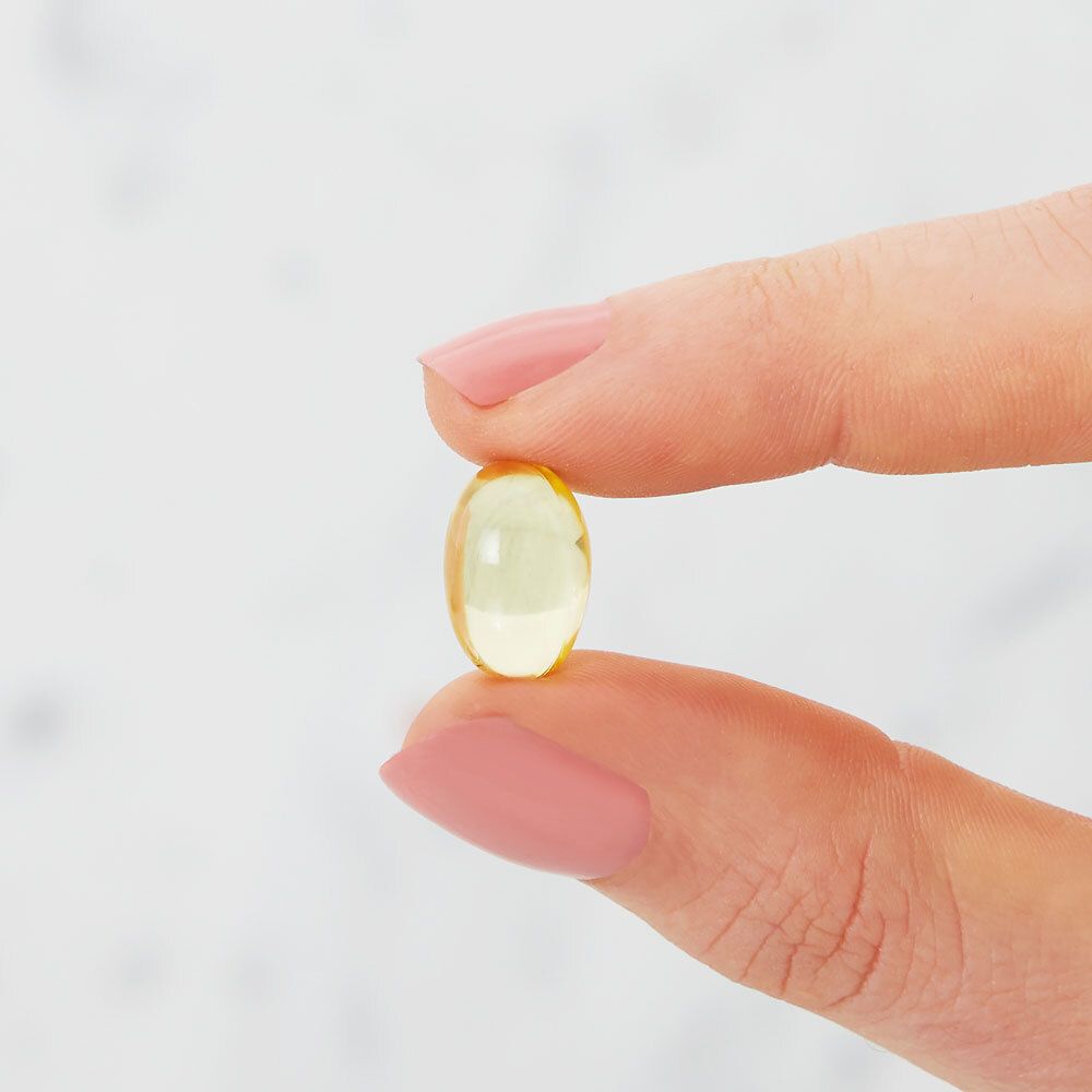 Gros plan d'une gélule molle jaune ovale, tenue entre les doigts. Fond clair et flou.