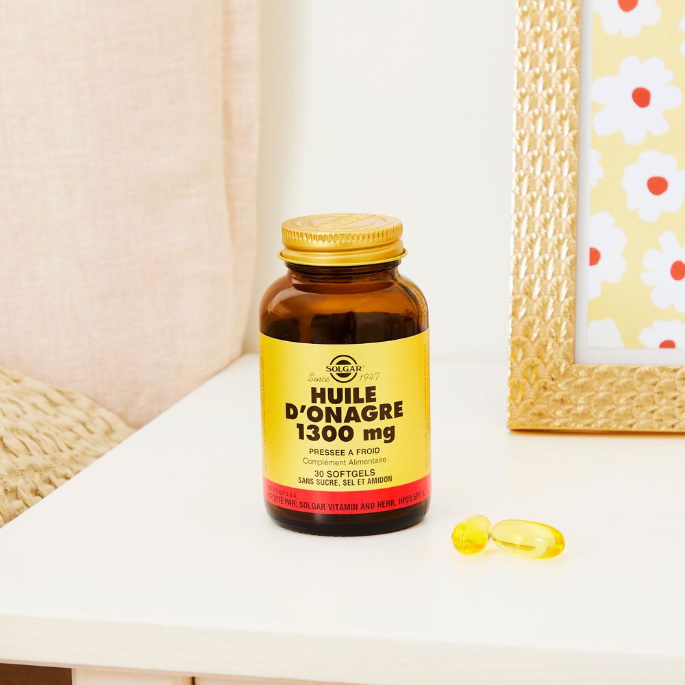 Flacon brun, bouchon doré. Deux gélules à côté. Sur une table, près d'un cadre photo.