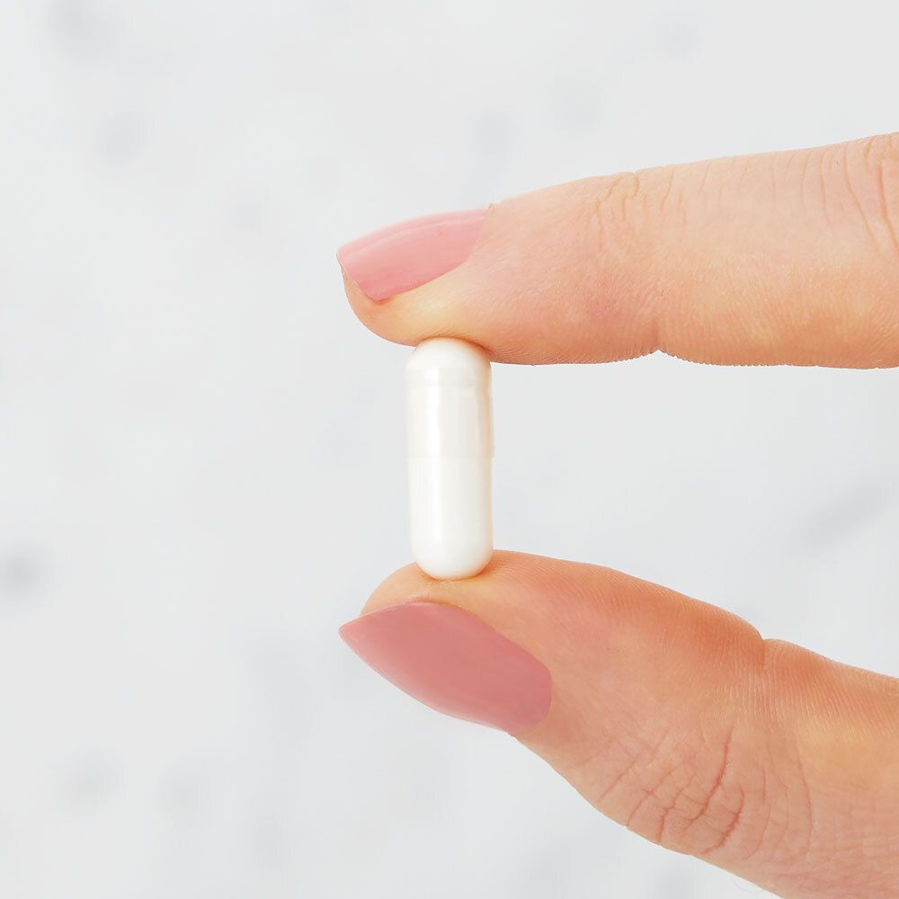 Une gélule blanche entre deux doigts. Ongles vernis roses.