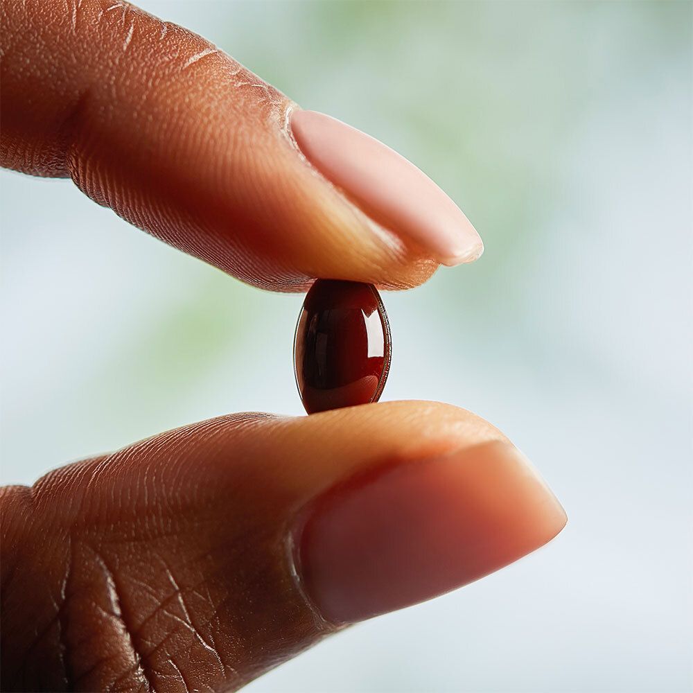 Une gélule rouge foncé tenue entre deux doigts.
