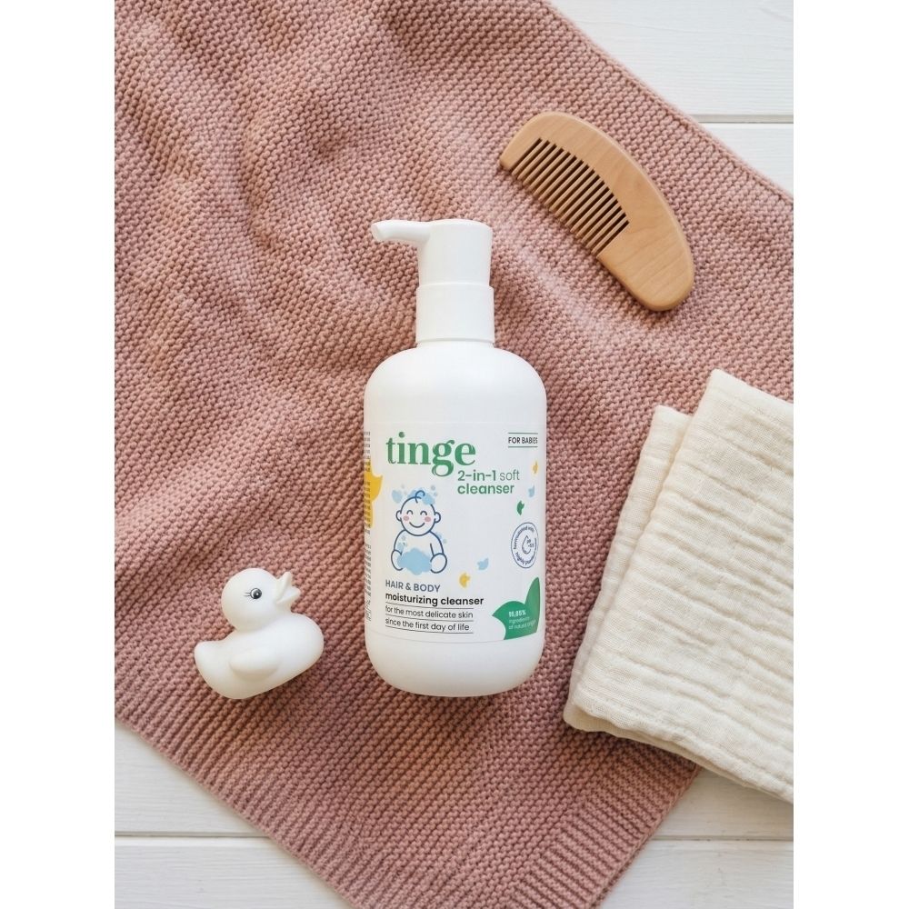 Flacon blanc avec pompe. Inscription : Tinge for babies, gel nettoyant 2 en 1. À côté d'une peluche et d'une serviette.