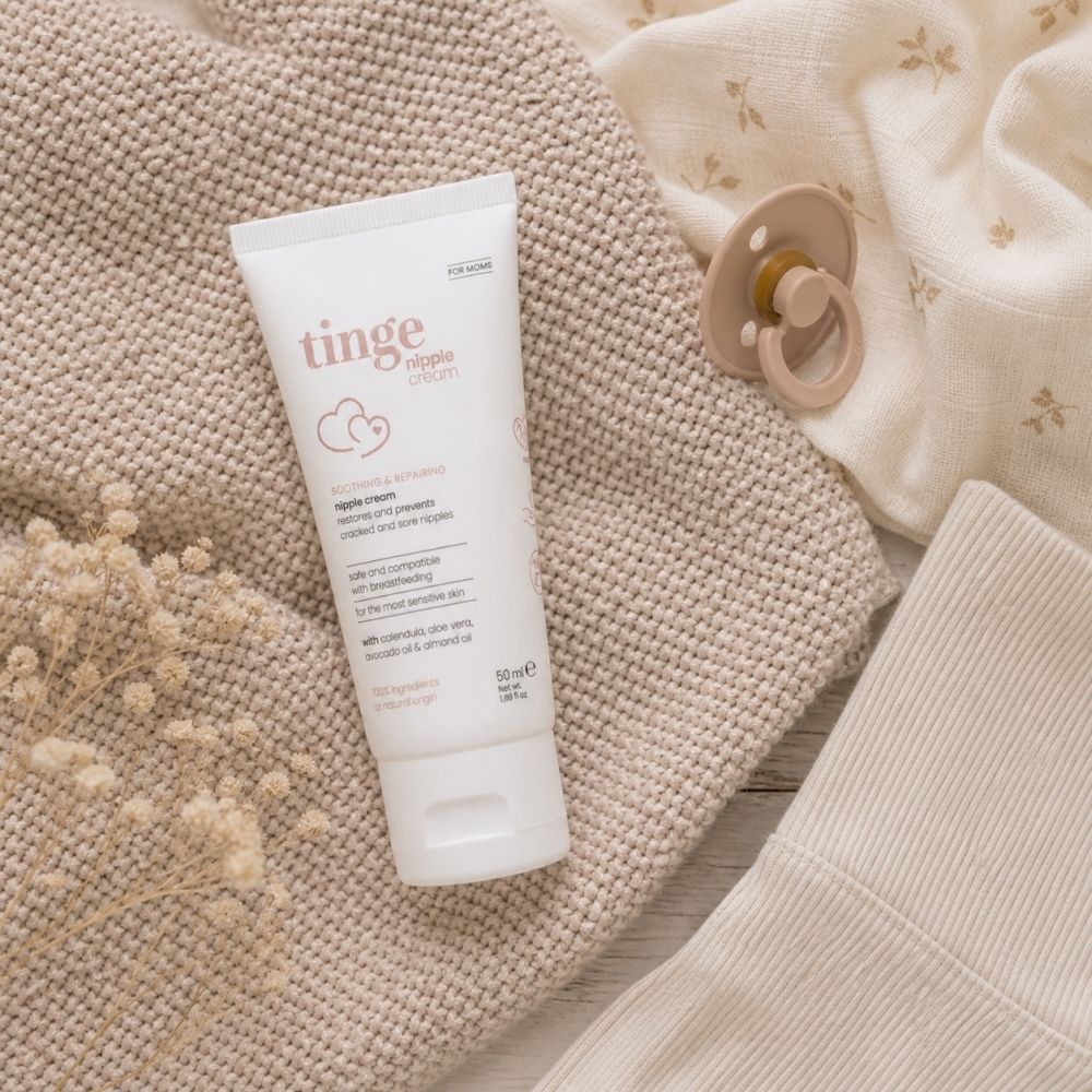 Tube de crème pour mamelons Tinge for Moms, 50 ml, drapé sur un tissu doux, à côté d'une sucette.