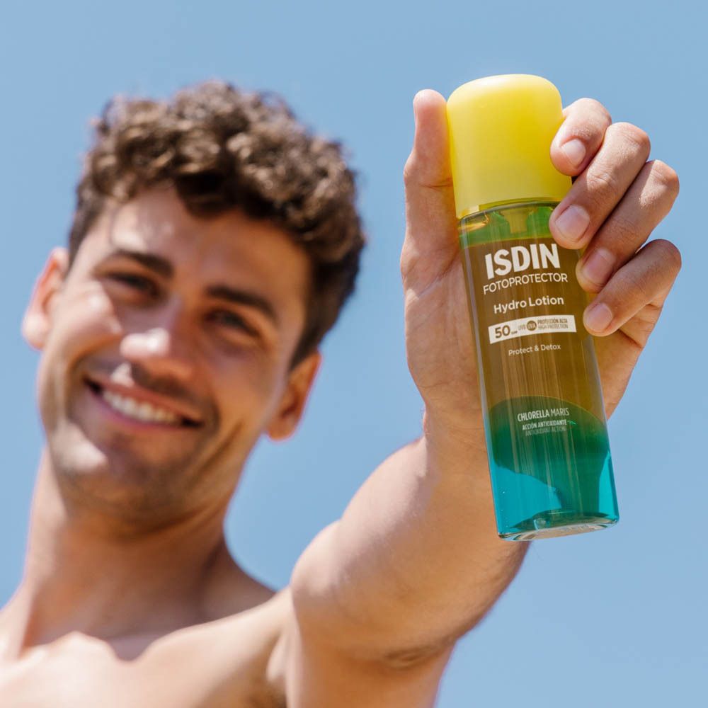 Flacon ISDIN Hydro Lotion SPF 50+. Liquide jaune transparent. Homme tenant le flacon. Ciel bleu.