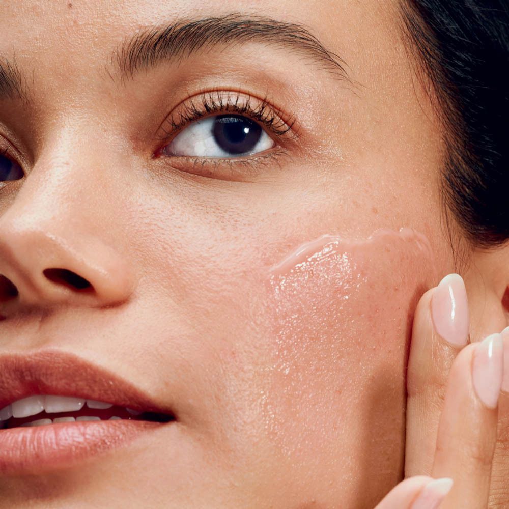 Gros plan d'une femme appliquant la crème hydratante CLINIQUE Moisture Surge sur sa joue.