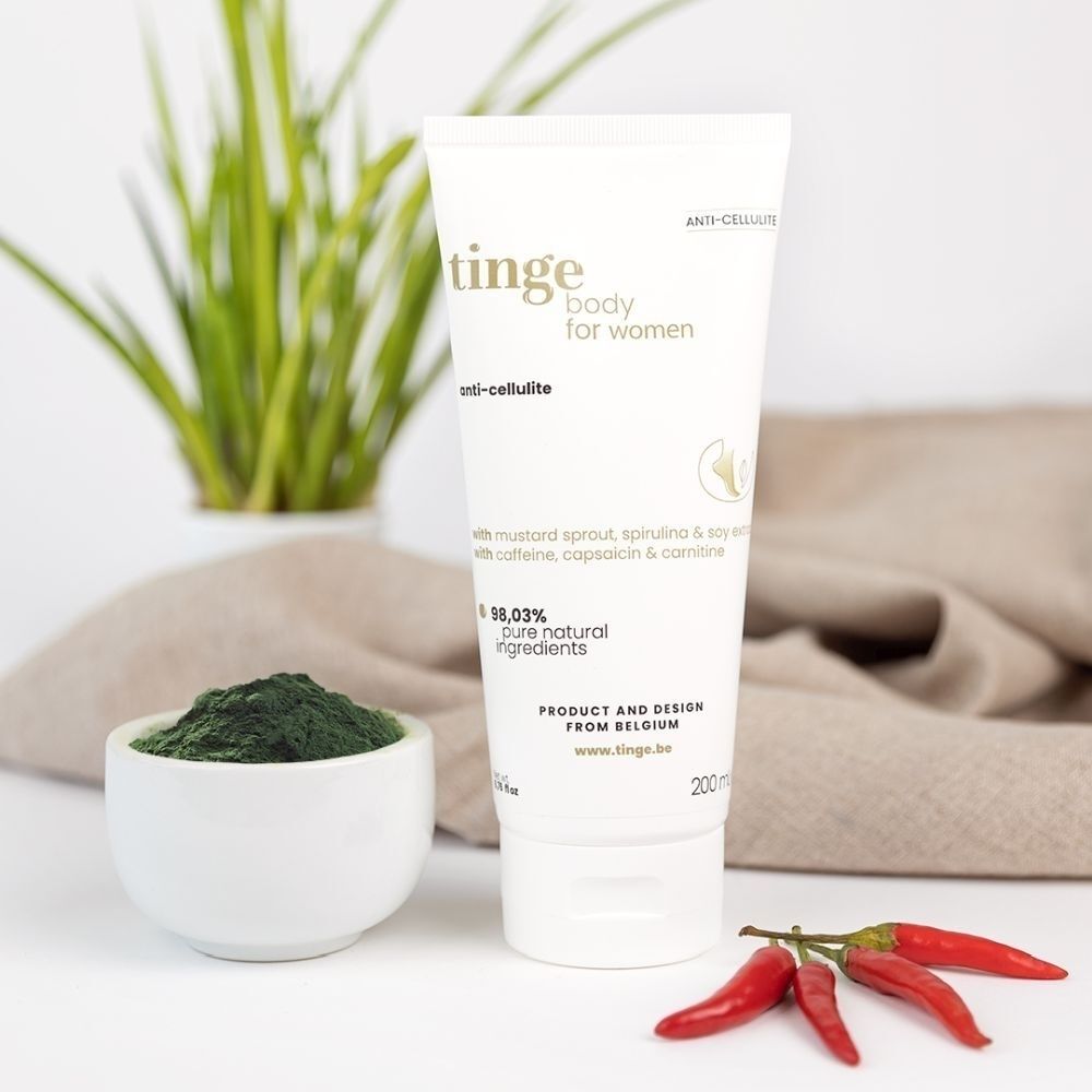 Tube de crème anti-cellulite Tinge avec poudre de spiruline et piments, entouré de plantes et de tissu.