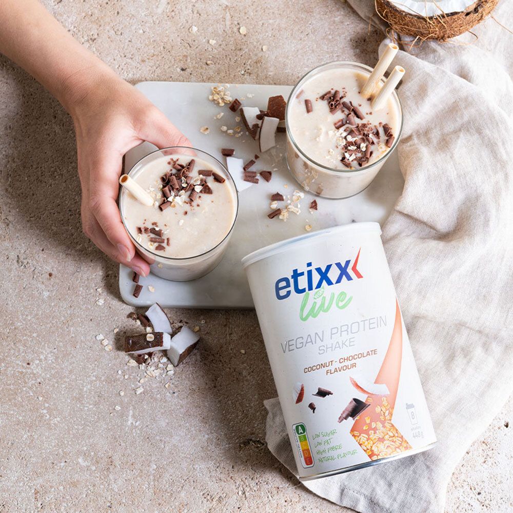 Deux verres de etixx Live Vegan Protein Shake. Garniture noix de coco et chocolat. Boîte à côté. Main tenant un verre.