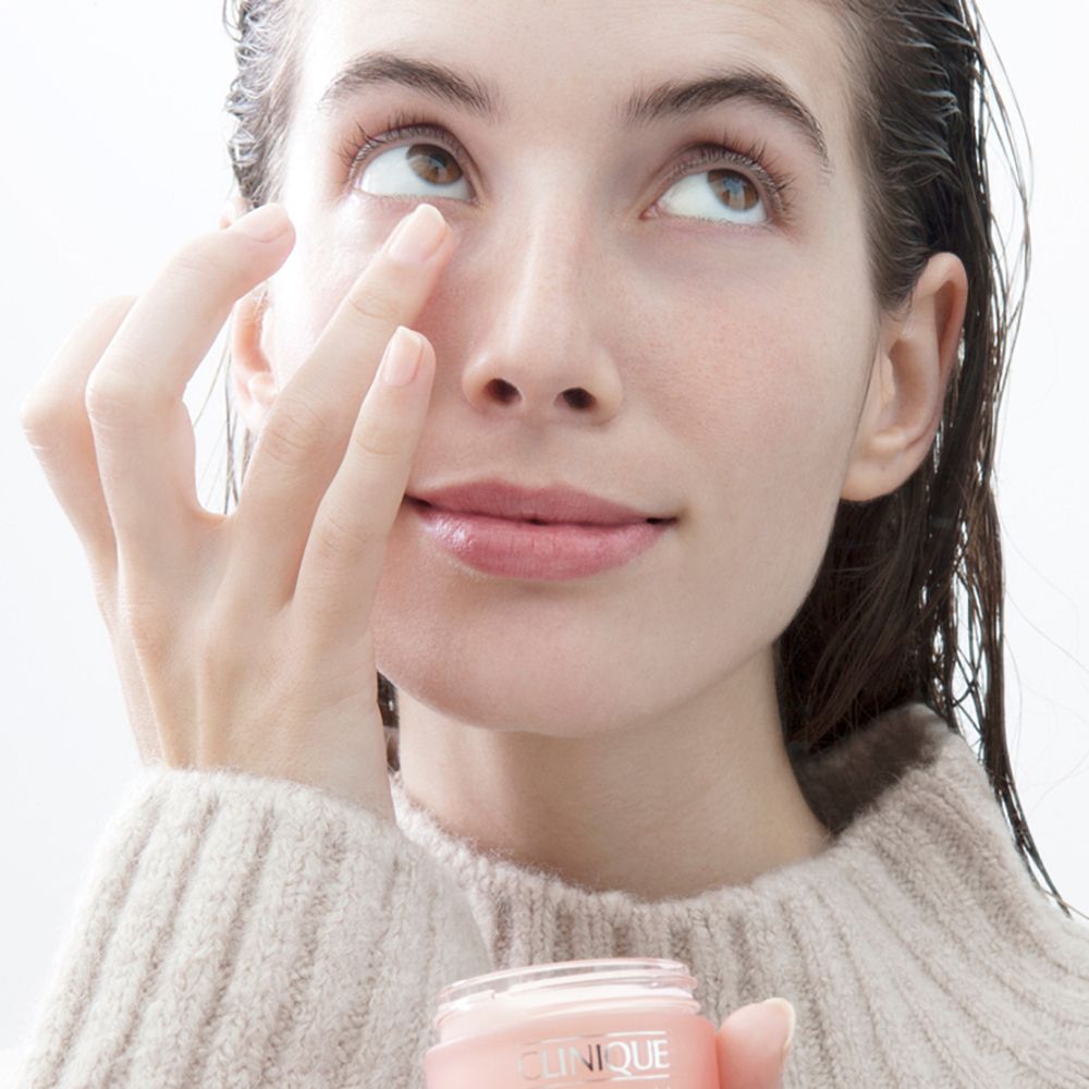 Femme tenant un pot de crème rose CLINIQUE. Elle touche le contour de ses yeux. Arrière-plan : clair.