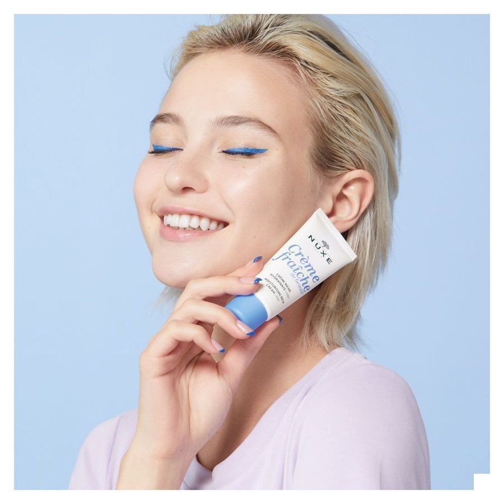 Femme tenant un tube Nuxe Crème Fraîche de Beauté contre sa joue, fond bleu.