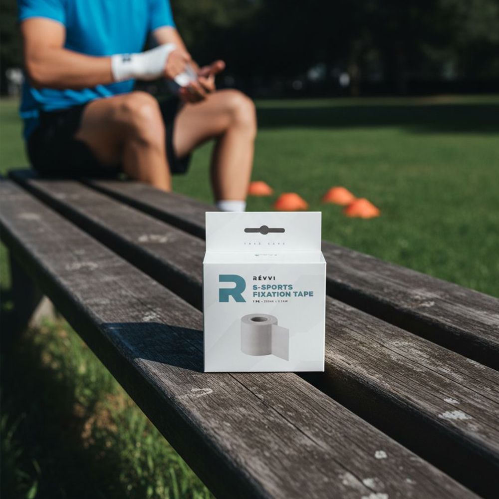 Emballage avec rouleau de ruban adhésif. Marque Révvi. Texte: S-SPORTS FIXATION TAPE, 1 pièce, 50 mm x 9,14 m. Sur banc.