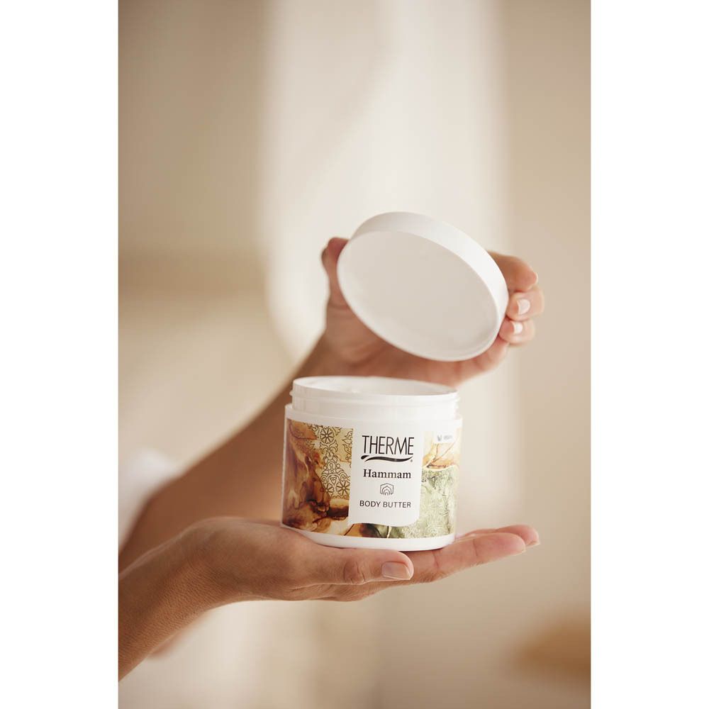 Mains tenant un pot blanc avec THERME Hammam BODY BUTTER. Couvercle à côté.