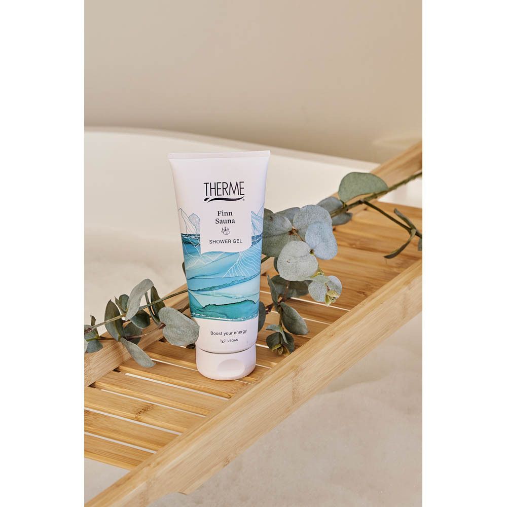 Tube de gel douche THERME Finn Sauna sur un plateau de baignoire en bois avec des branches de plantes.
