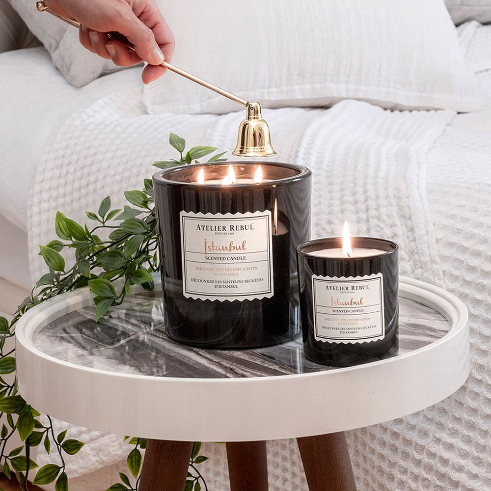 Deux bougies noires allumées. Étiquette avec texte: Atelier Rebul Istanbul Scented Candle. Cloche dorée.