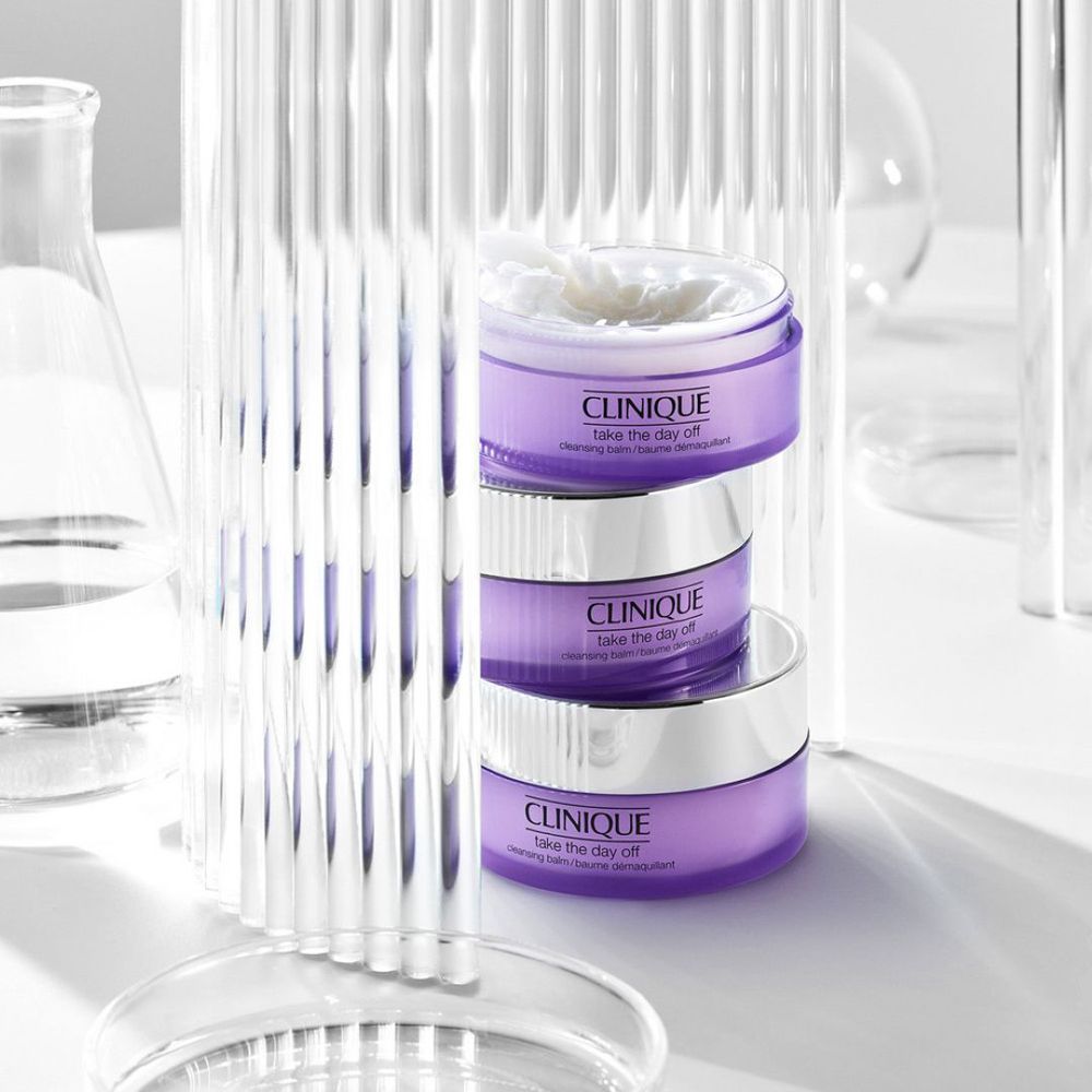 Trois pots CLINIQUE Take the Day Off, empilés. Verrerie et matériel de laboratoire en arrière-plan.