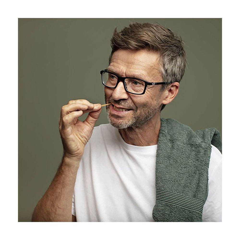 Un homme utilise un cure-dent. Il porte des lunettes et un t-shirt blanc. Une serviette est sur son épaule.