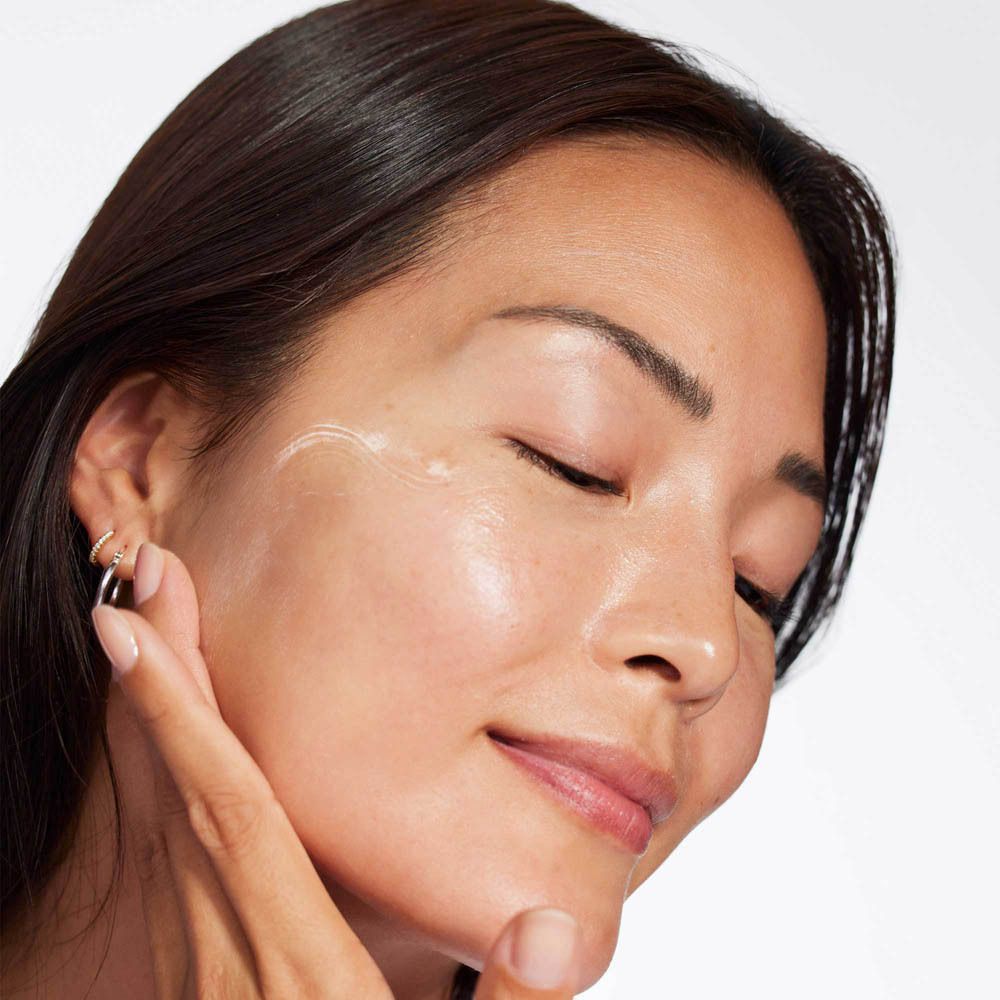 Femme aux yeux fermés appliquant une crème blanche sur sa joue.