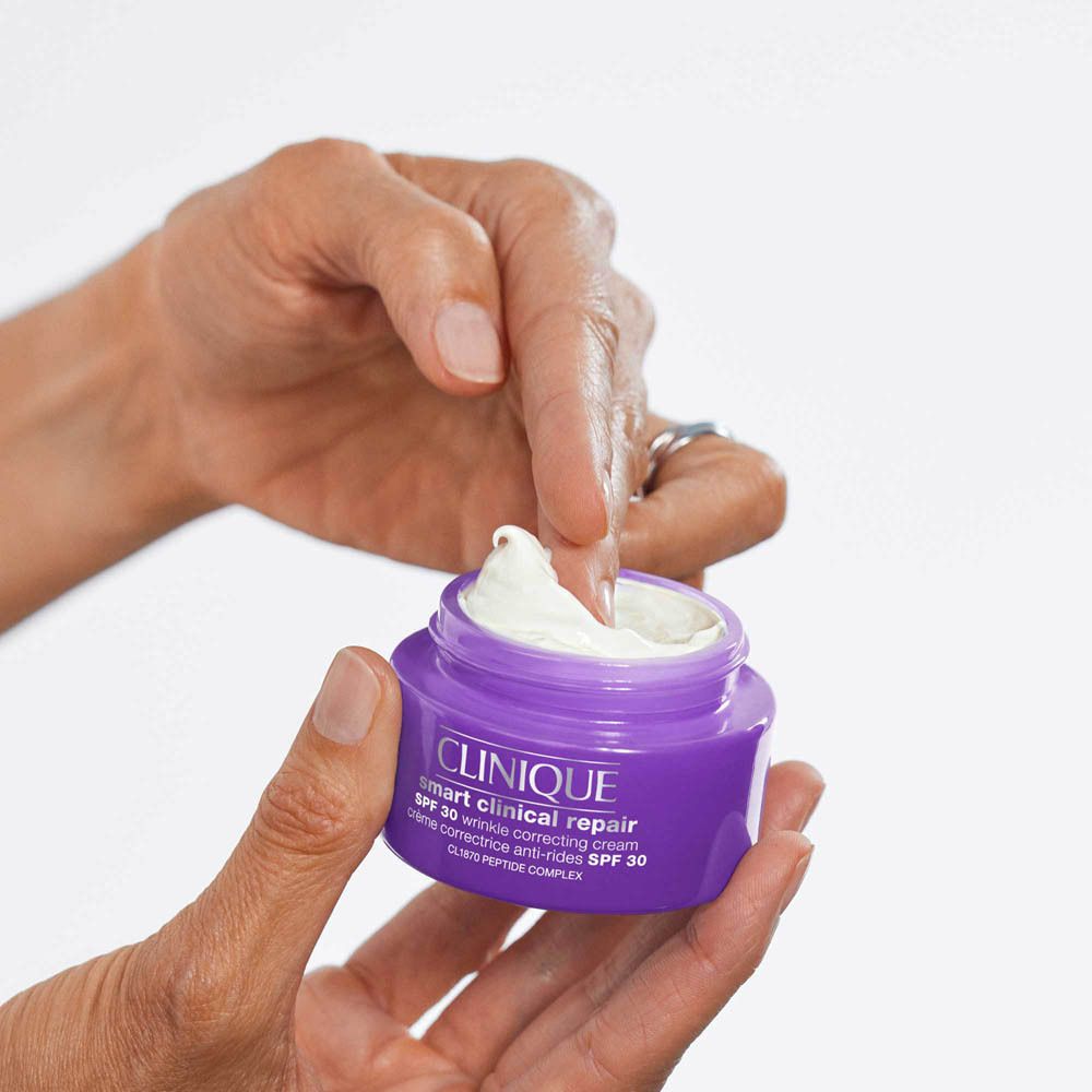 Une main ouvre un pot violet de crème hydratante Clinique Smart Clinical Repair SPF 30.