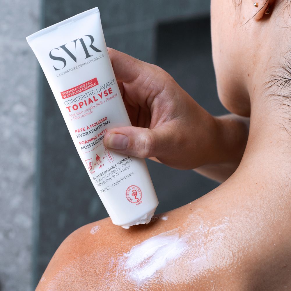Personne appliquant le produit sur l'épaule. Crème blanche. Tube à la main. Arrière-plan : salle de bain.