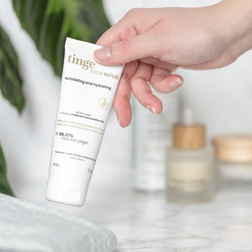 Main tenant un tube Tinge Gommage Visage devant des bouteilles floues et des plantes. Origine naturelle 98,37%.