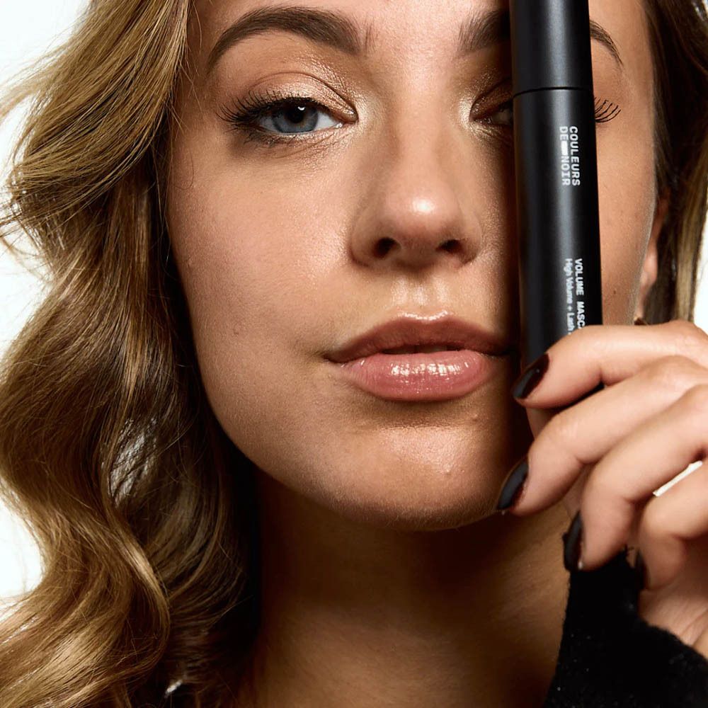 Femme tenant un tube de mascara. Texte: Couleurs de Noir, Volume Mascara, High Volume + Lash Boost.