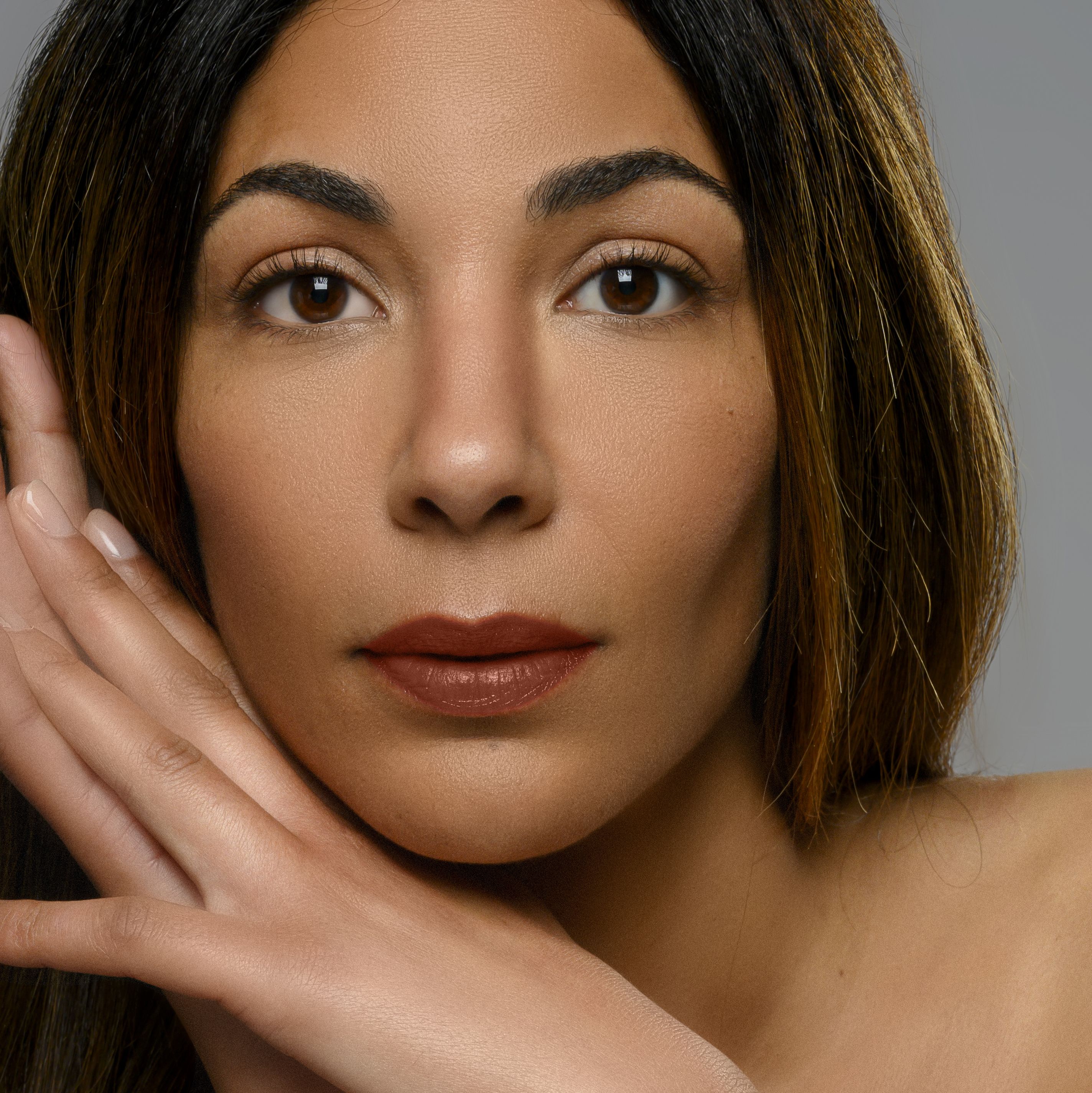 Portrait d'une femme portant du rouge à lèvres. Lèvres brun-rouge, sourcils et cheveux foncés. Mains sur le visage.
