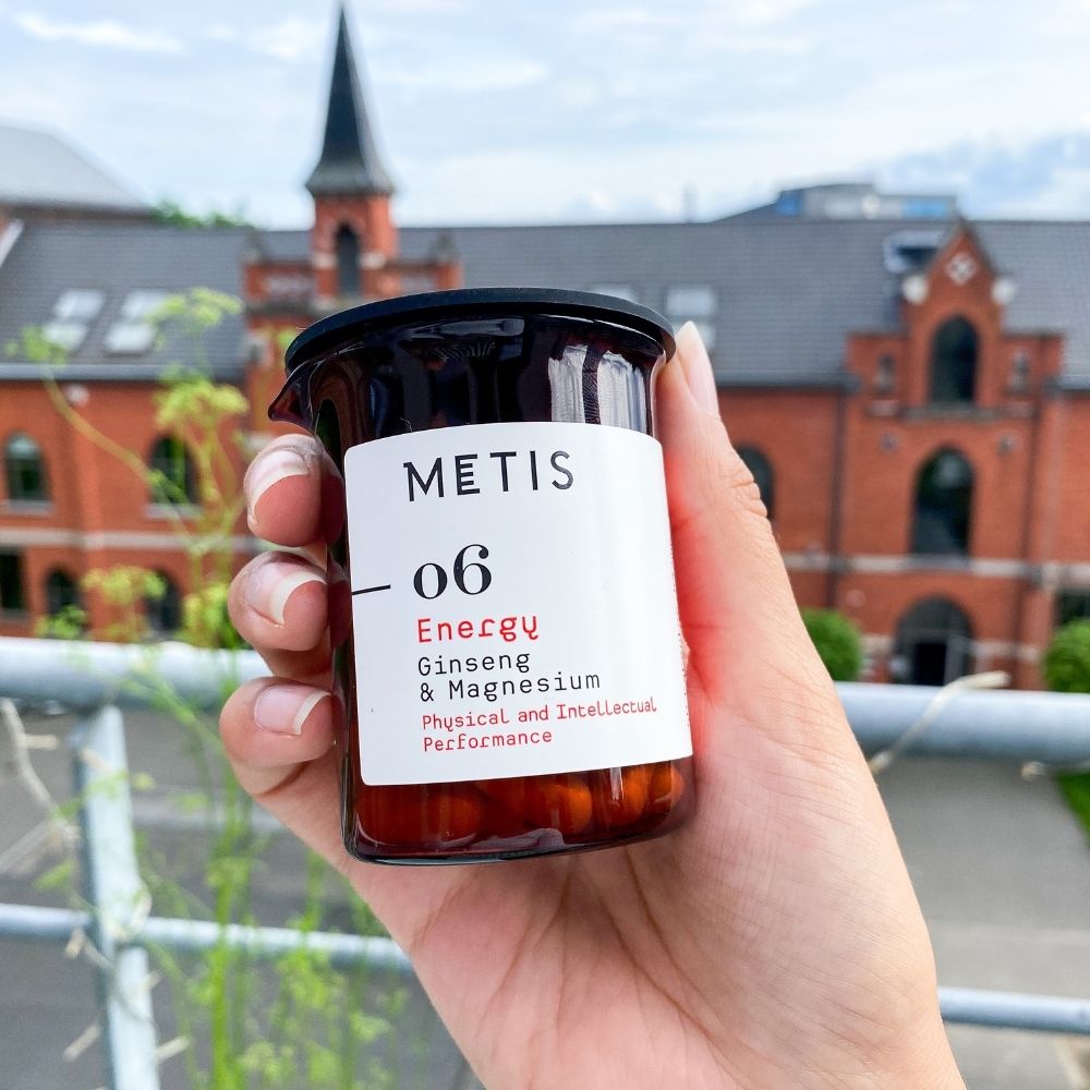 Main tenant un bocal marron avec Metis Energy 06. Gélules rouges visibles. Fond: bâtiment.