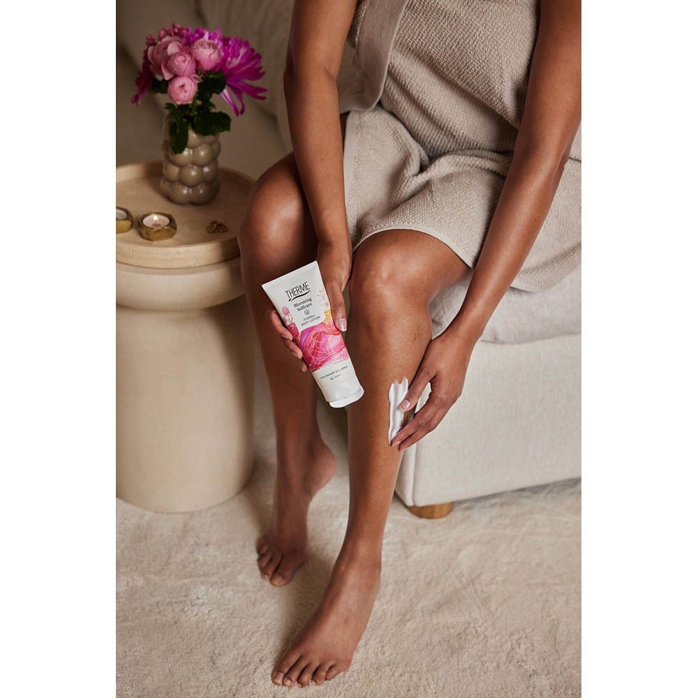 Personne appliquant THERME Blooming Selfcare Body Lotion sur la jambe. Design floral, texte.