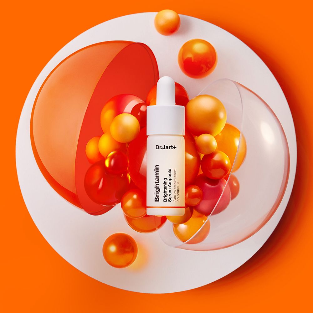 Petite bouteille avec pipette sur une assiette blanche avec des billes oranges. Inscription : Dr.Jart+ Brightamin Brightening Serum Ampoule.