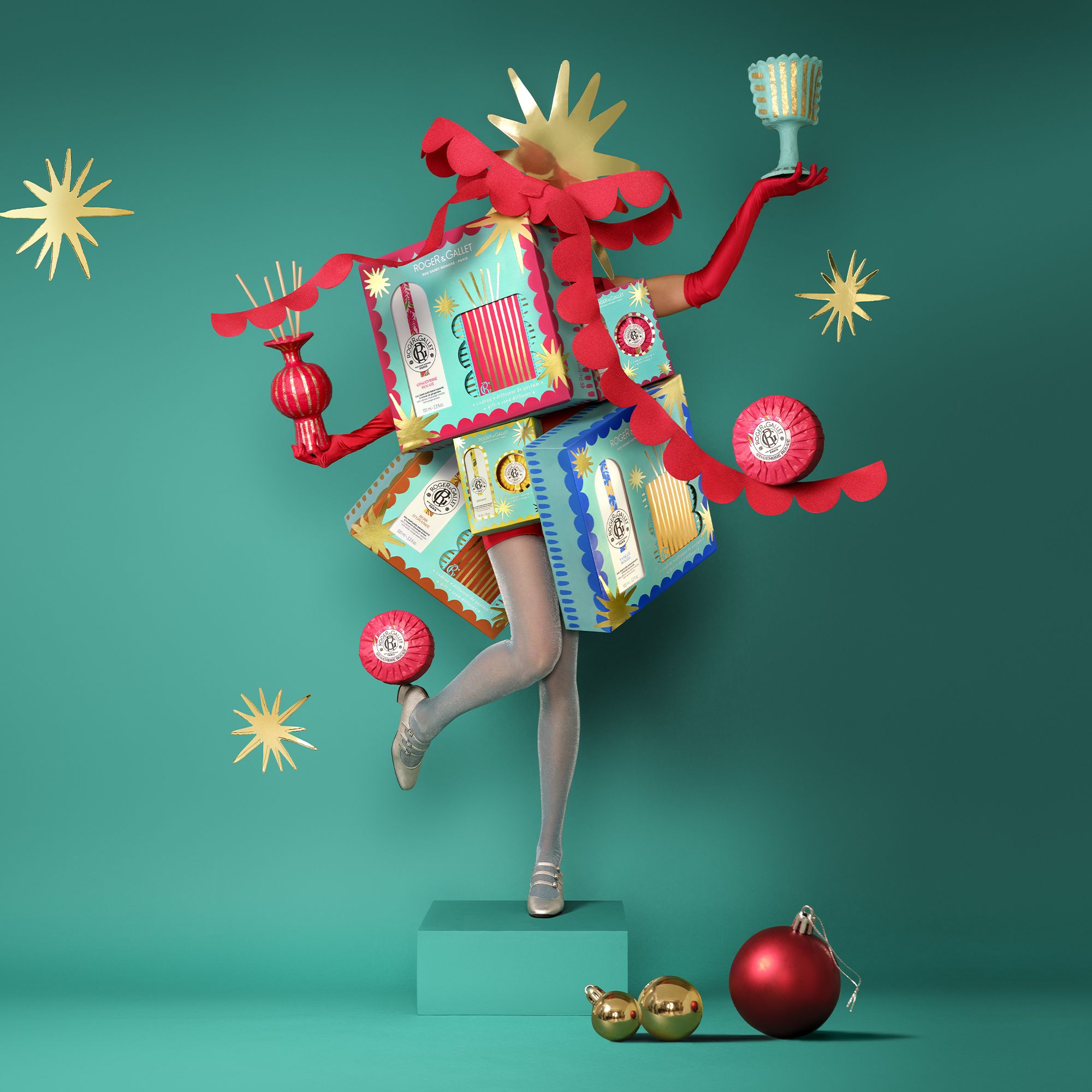 Mise en scène créative. Personne avec coffrets cadeaux. Produits Roger & Gallet. Décoration avec étoiles et boules.