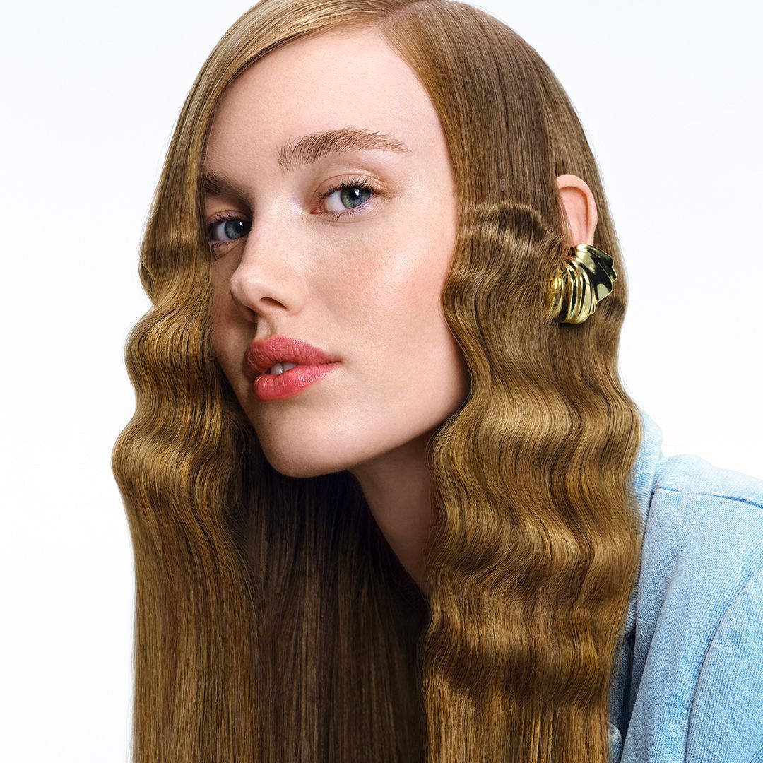 Portrait d'une femme aux cheveux ondulés. Elle porte une boucle d'oreille dorée. Peau claire, lèvres roses.