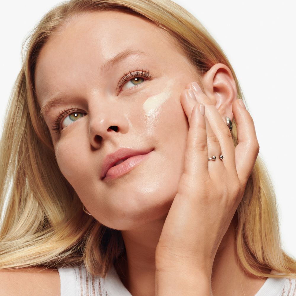 Femme appliquant de la crème sur son visage. Anneaux aux doigts. Peau claire, cheveux blonds.
