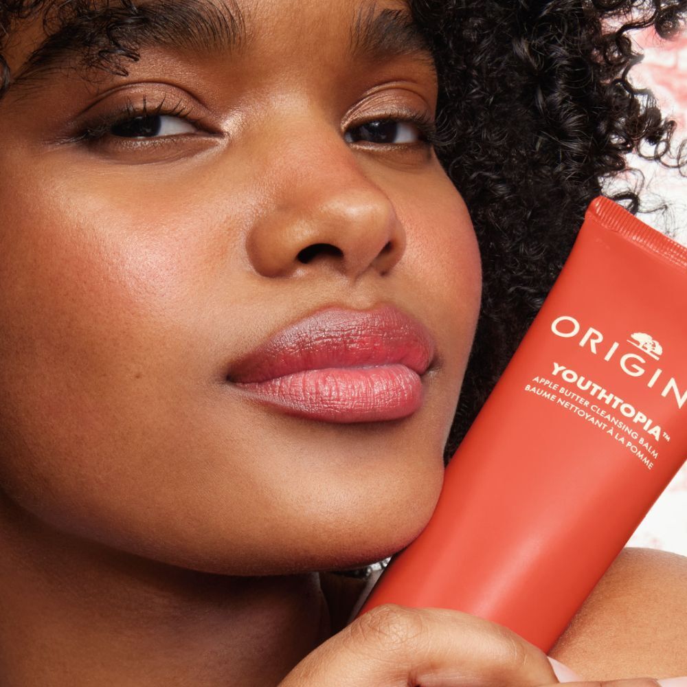 Femme tenant un tube rouge. Origins Youthtopia Apple Butter Cleansing Balm est visible.