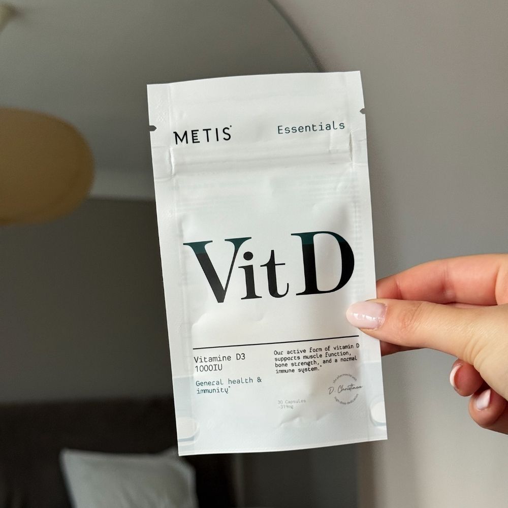 Main tenant un emballage de Metis Essentials Vit D. Inscription : Vit D, vitamine D3 1000UI.