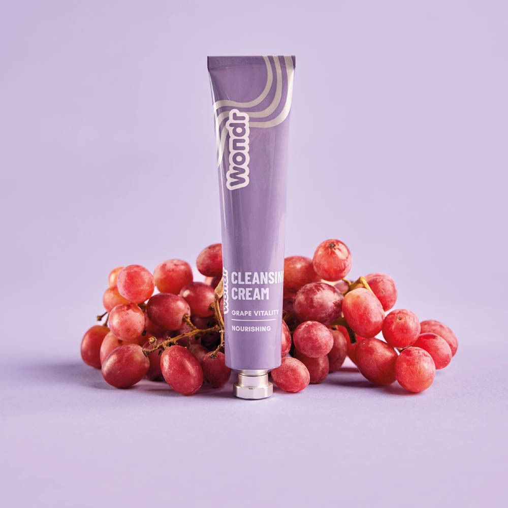 Tube violet avec logo "Wondr" et texte "Cleansing Cream Grape Vitality". Posé sur des raisins. Sur fond violet.