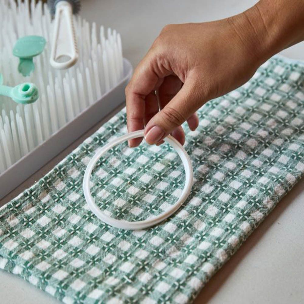 Une main tient un anneau blanc en silicone sur une serviette. Un égouttoir avec des accessoires pour biberons est visible en arrière-plan.