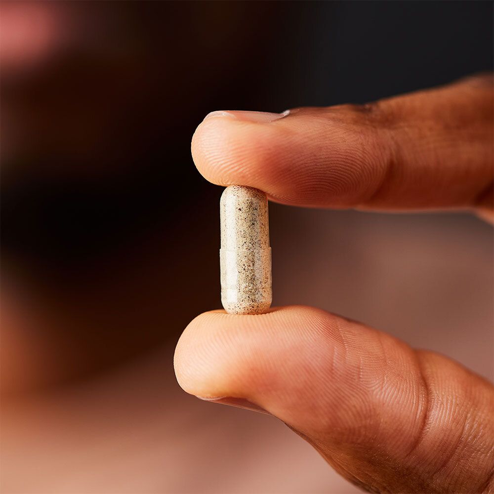 Une gélule tenue entre deux doigts. Beige avec des points foncés.