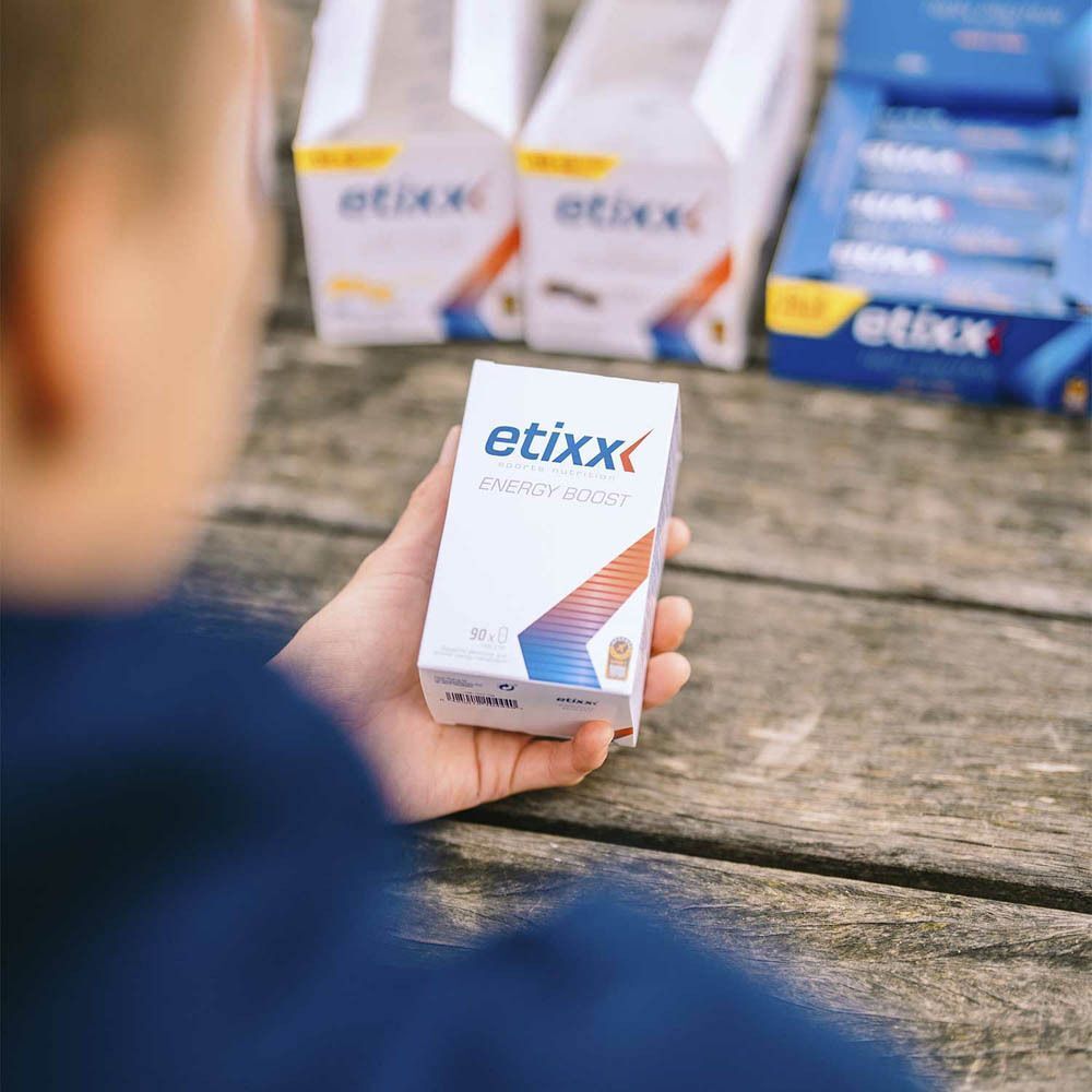 Personne tenant une boîte blanche Etixx Energy Boost. Autres boîtes en arrière-plan.
