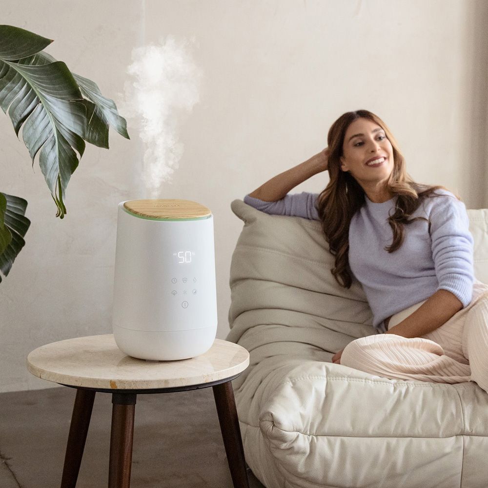 Humidificateur blanc avec couvercle en bois. Vapeur visible. Femme assise sur un canapé.