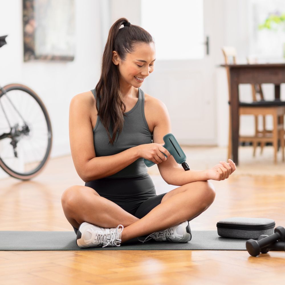 Une femme utilise un mini pistolet de massage vert. Marque : medisana. Le nom du produit et le logo sont visibles. Assise sur un tapis.