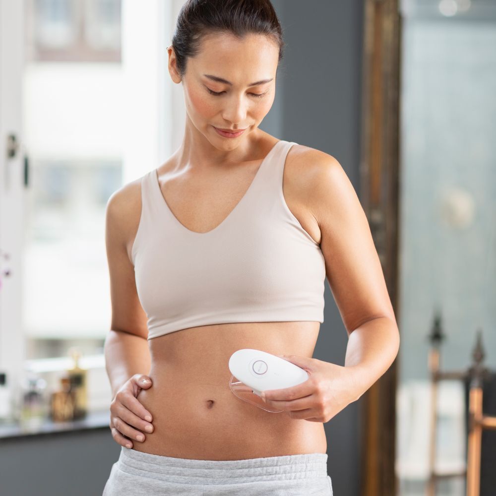 Femme tenant un appareil de massage blanc, medisana. Application sur l'abdomen. Porte un soutien-gorge et un pantalon de sport.