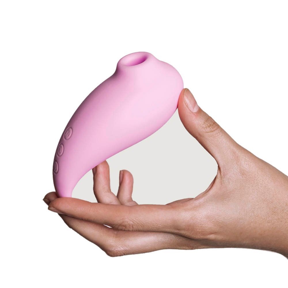 Stimulateur rose tenu dans une main. Forme ovale avec ouverture et boutons. Fond blanc.