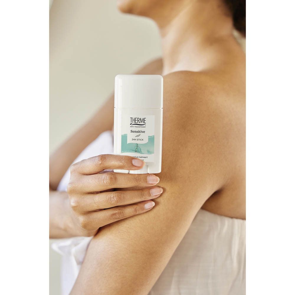 Personne tenant un stick anti-transpirant blanc sous le bras. Produit avec inscription THERME, Sensitive, 24H STICK.