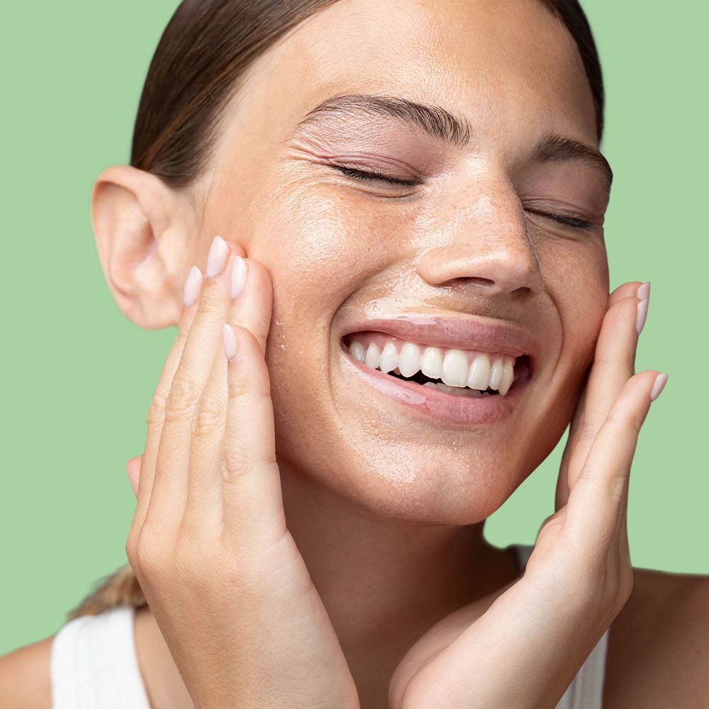 Femme aux yeux fermés, appliquant de l'huile sur son visage. Fond vert.