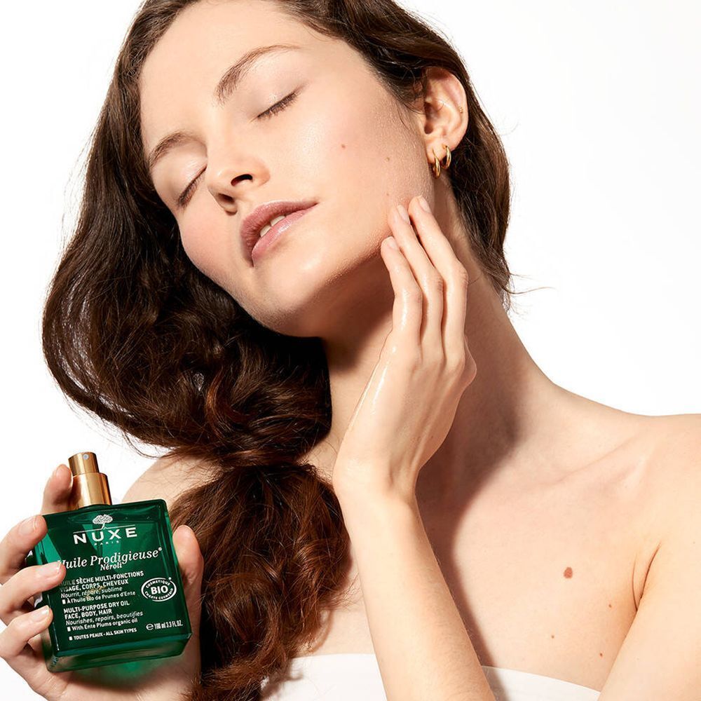 Femme appliquant de l'huile sur son visage. Flacon vert avec bouchon doré. Inscription : NUXE.