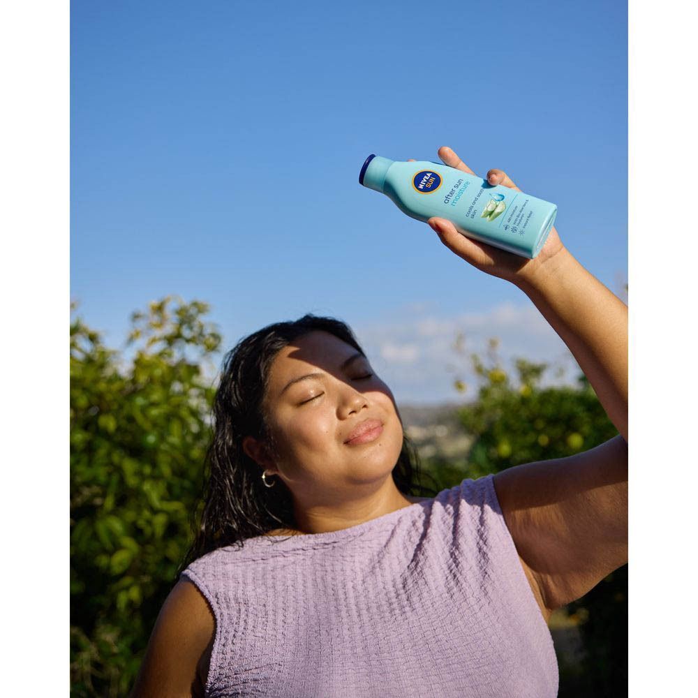 Une femme tient une bouteille bleue NIVEA SUN After Sun. Elle sourit. Fond : ciel bleu.
