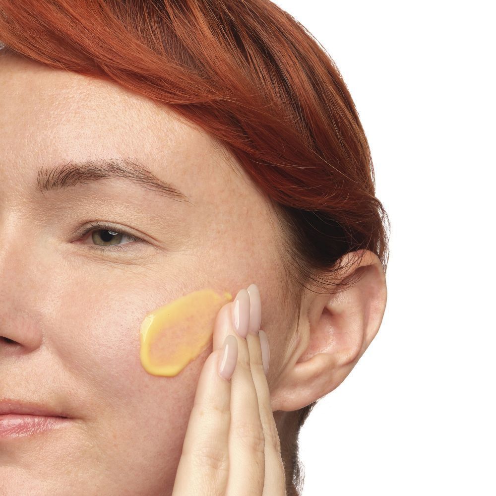 Une main applique une émulsion jaune sur la peau. Une partie du visage est visible.